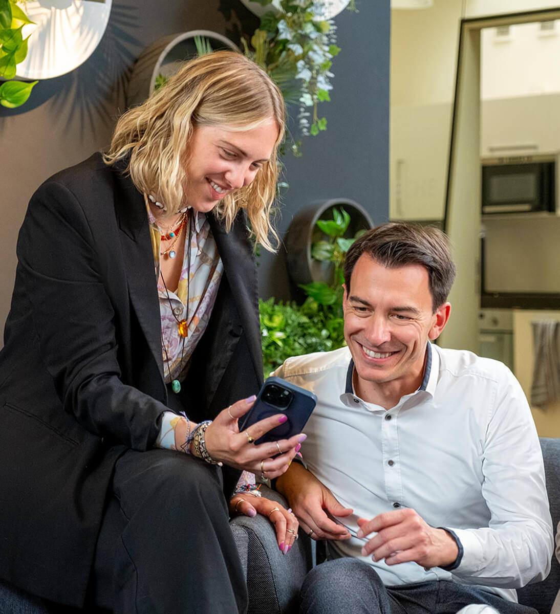 A young, blonde woman is leaning on the armchair, where the man is sittint. She is holding up a phone, so he can see what is on the screen. | google ads conversion audit SUNZINET